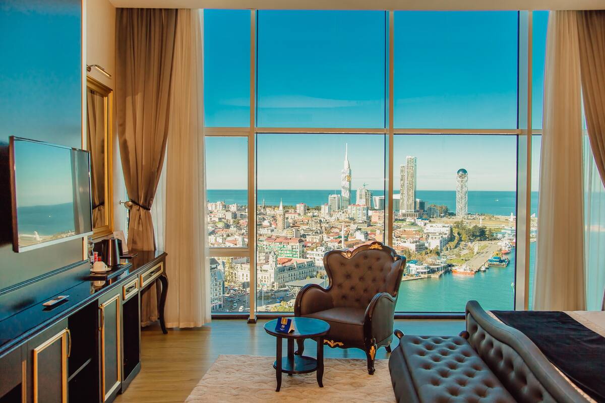 Ein Hotelzimmer mit großem Fenster und Blick auf die Stadt, einer Couch, einem kleinen Tisch und einem Fernseher auf einem Schrank.