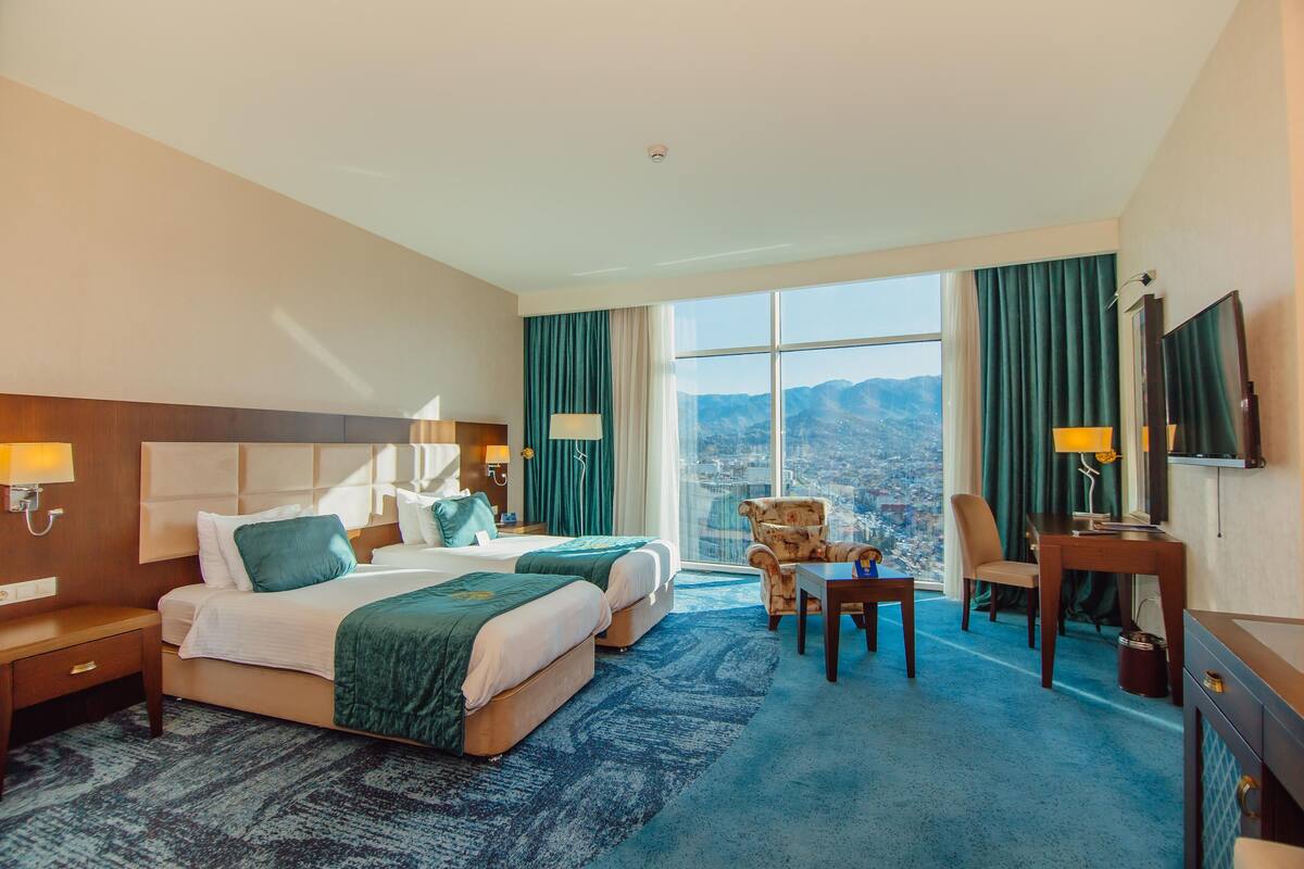 Ein Hotelzimmer mit zwei Betten, einem Schreibtisch, einem Sessel, einem Fernseher und einem großen Fenster mit Blick auf die Berge.