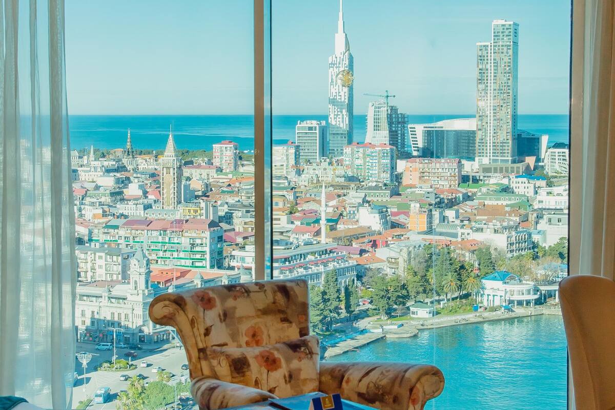 Ein Zimmer mit großem Fenster und Blick auf die Stadt, mit einem floralen Sessel, einem kleinen Tisch und einem blauen Teppich.