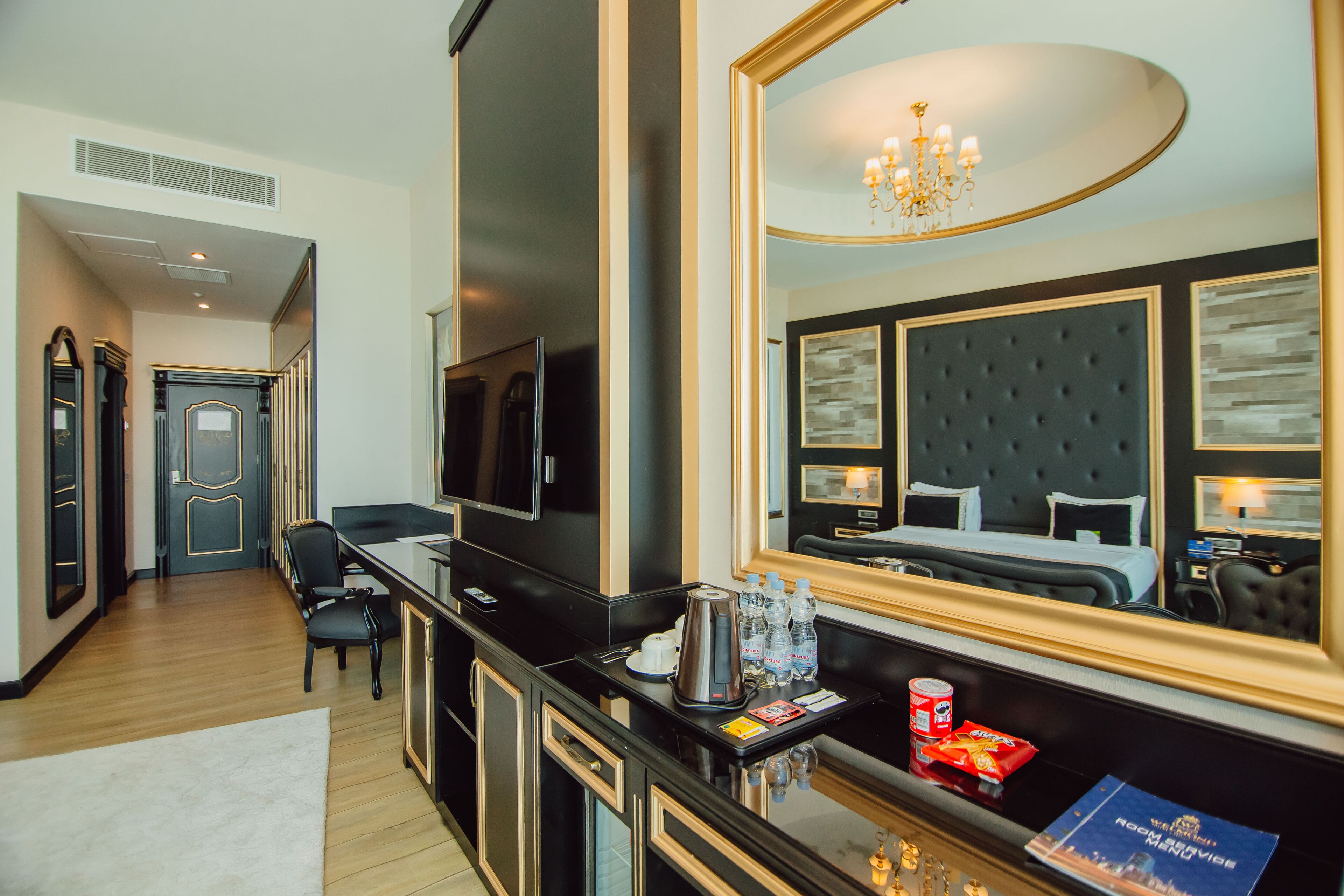 A modern hotel room with a large mirror, a flat-screen TV, a minibar, and a comfortable bed.