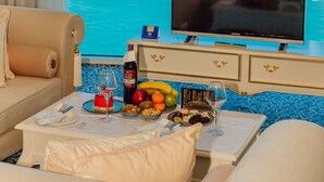 A hotel room with a large TV, a dining table with food and drinks, a chandelier, and a view of the sea. - JRW Welmond Hotel & Casino Batumi (Batumi)