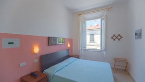 Une chambre à coucher comprenant un lit, une table de chevet, une lampe fixée au mur, une fenêtre et un petit banc.