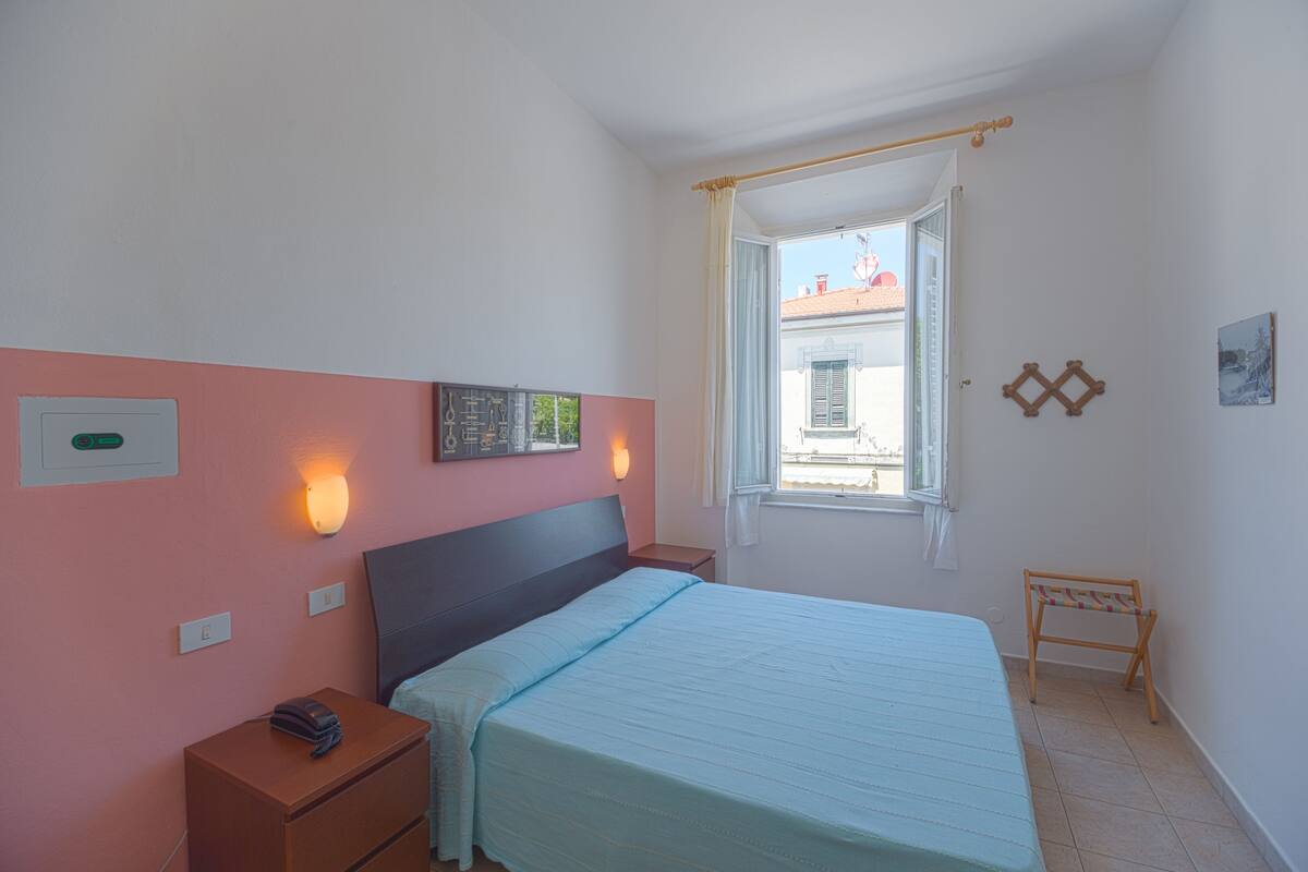 Een slaapkamer met een bed, nachtkastje, een wandlamp, raam en een klein bankje.