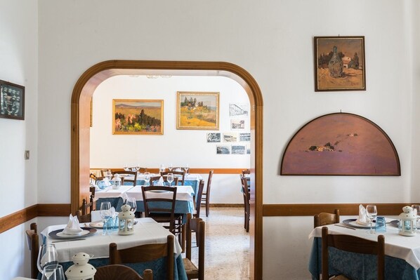 A dining area with tables set for two, framed artwork on the walls, and a view through an archway. - Hotel Fiammetta (Livorno)