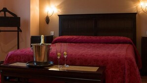 Una habitación de hotel con cama, mesita de noche con lámpara, otra mesita auxiliar, una lámpara de pared y un termostato.