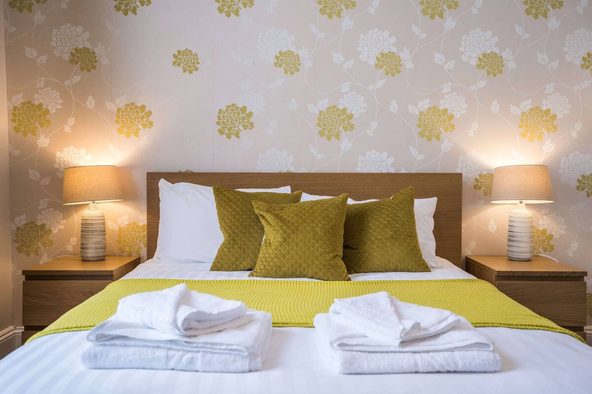 Un lit bien fait, avec du linge de lit blanc et des coussins verts, deux lampes de chevet et un papier peint à motifs floraux.
