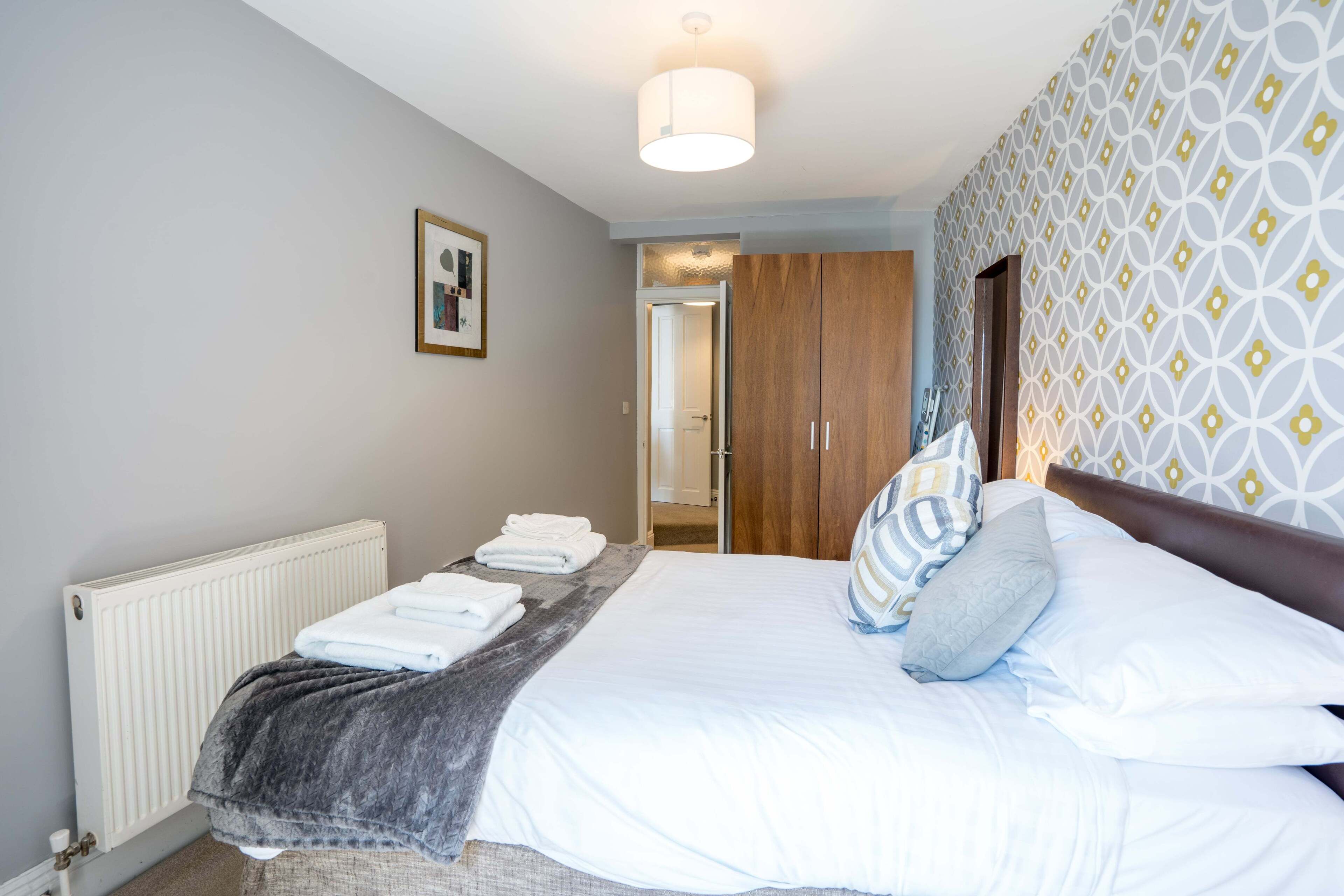 A neatly made bed with white linens and a grey blanket, a wooden wardrobe, and a patterned wall.