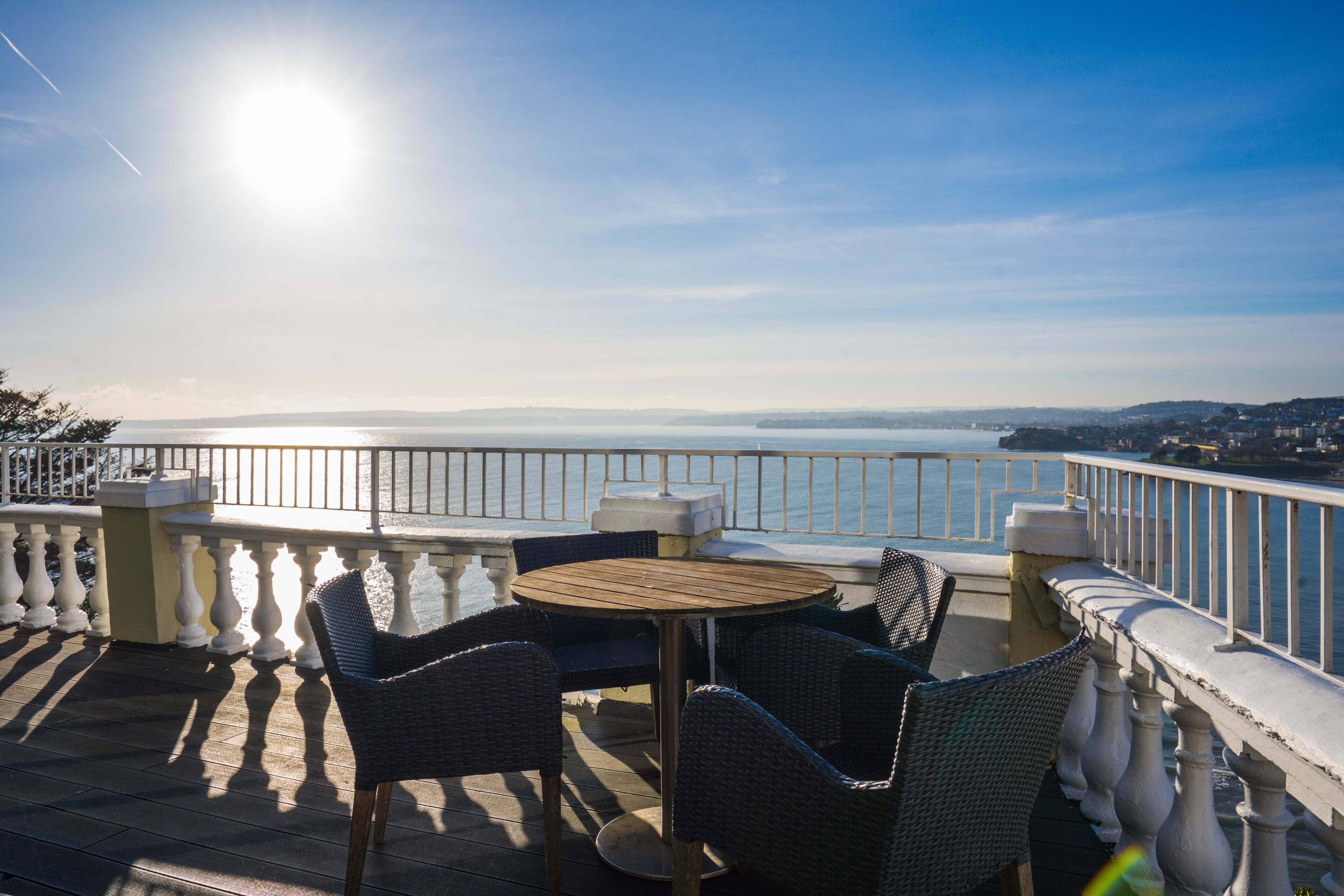 Ein Balkon mit Korbstühlen und einem runden Tisch, mit Blick auf ein Gewässer und das entfernte Land.
