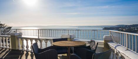 A balcony with wicker chairs and a round table, overlooking a body of water and distant land.