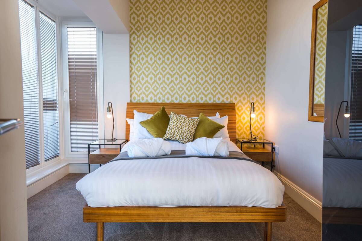 Un lit bien fait, avec du linge de lit blanc et des coussins verts, une table de chevet avec une lampe, et un papier peint à motifs sur le mur.