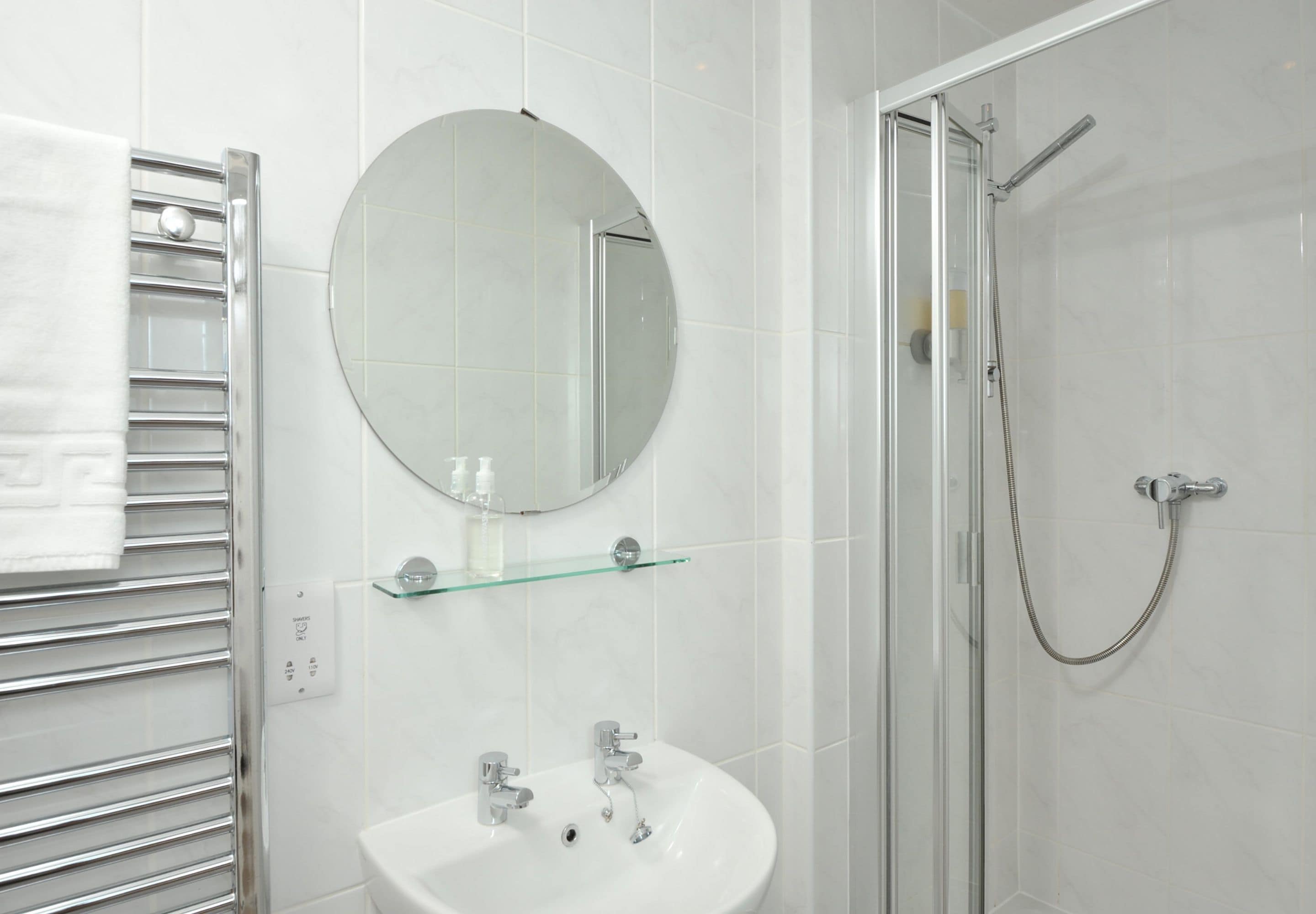 Un baño moderno con cabina de ducha de vidrio, un espejo redondo, un lavabo blanco y un perchero.