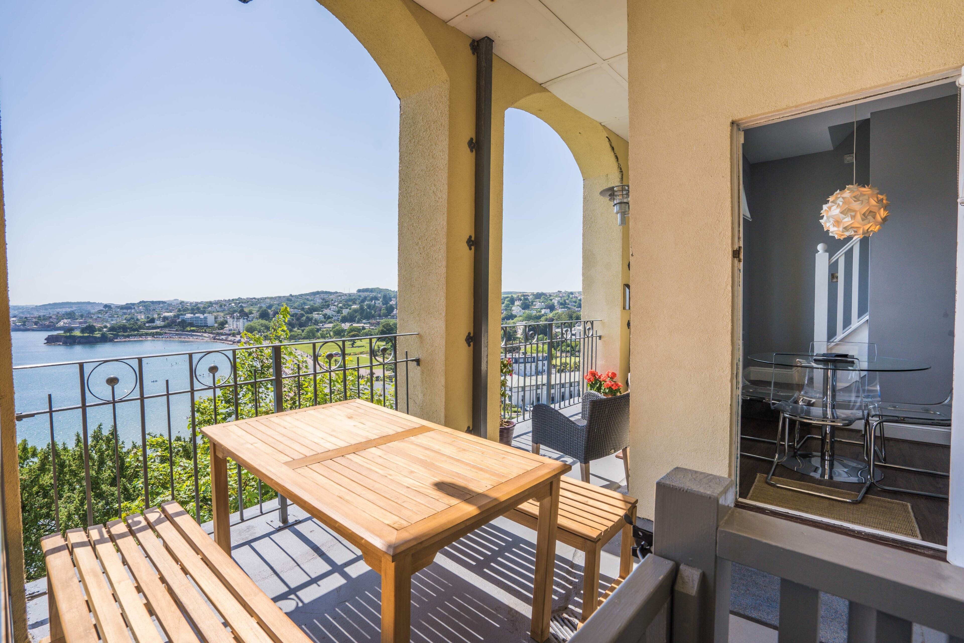 A balcony with a wooden table and chairs, overlooking a body of water and a cityscape.