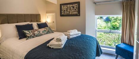 A neatly made bed with pillows, a blue quilt, and rolled towels. There is a window with a view of greenery outside.