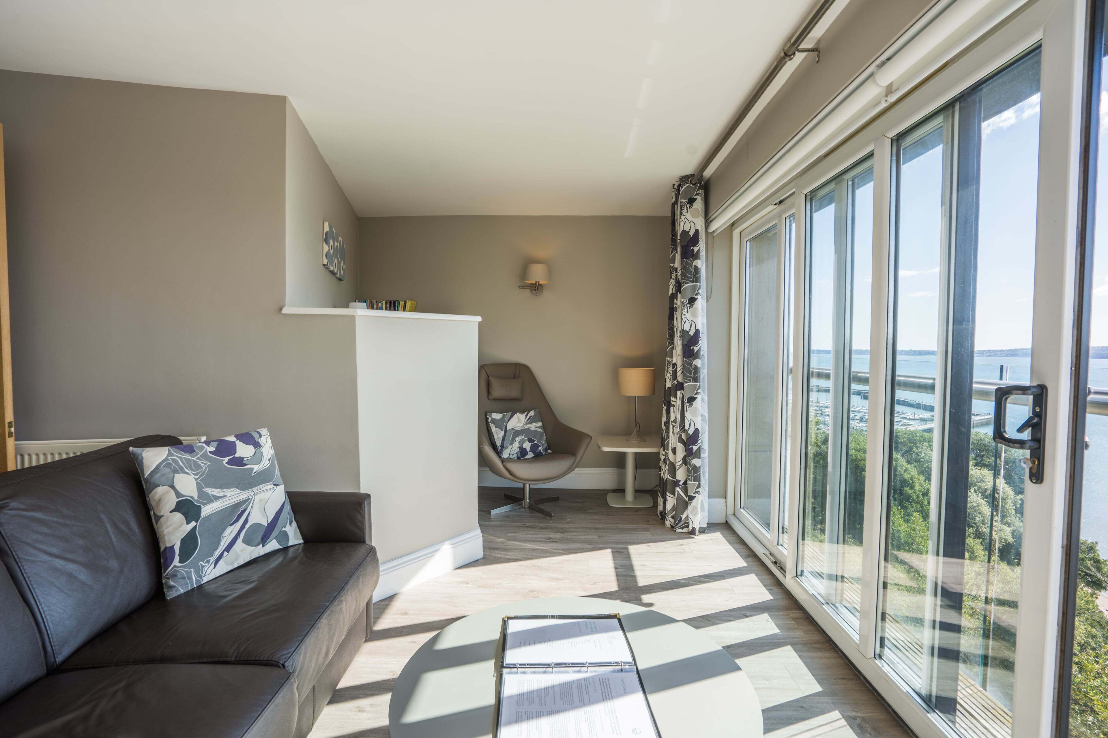 Ein modernes Wohnzimmer mit einer Couch, einem Sessel, einem Tisch und einem großen Fenster mit Meerblick.
