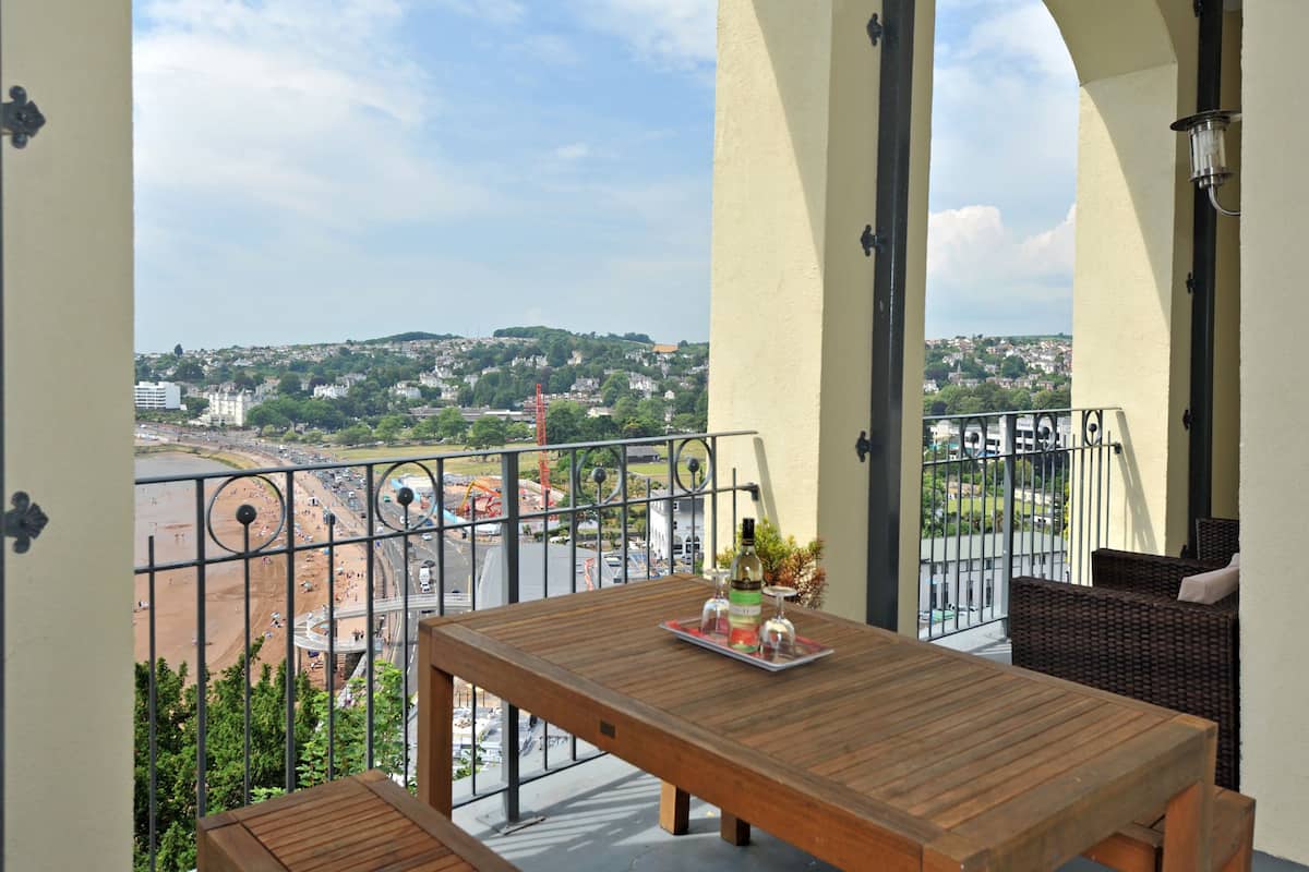Un balcon avec une table en bois, des meubles en osier et une vue sur la plage et le paysage urbain.
