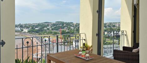 A balcony with a wooden table, wicker furniture, and a view of a beach and cityscape.