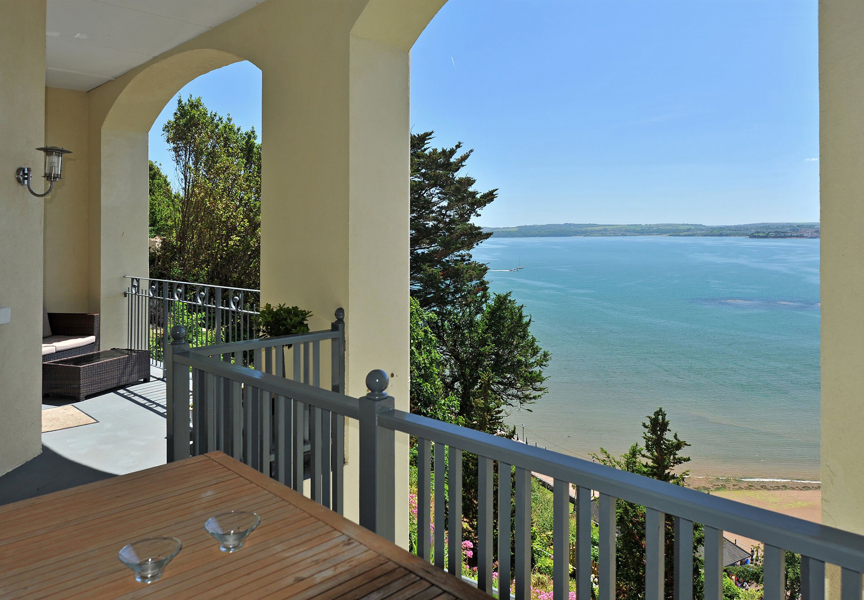 A balcony with a wooden table and two glasses, overlooking a calm sea and greenery.