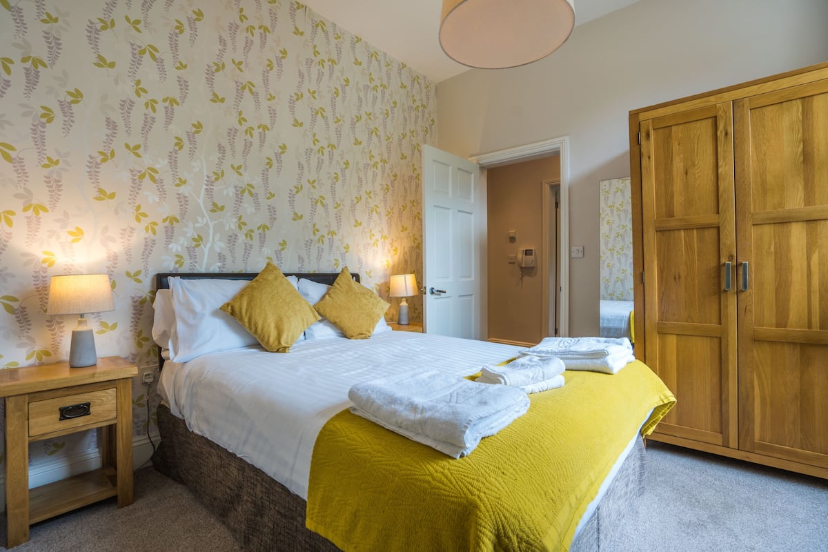Un lit bien fait, avec du linge de lit blanc et un plaid jaune, une armoire en bois et une table de chevet avec une lampe.