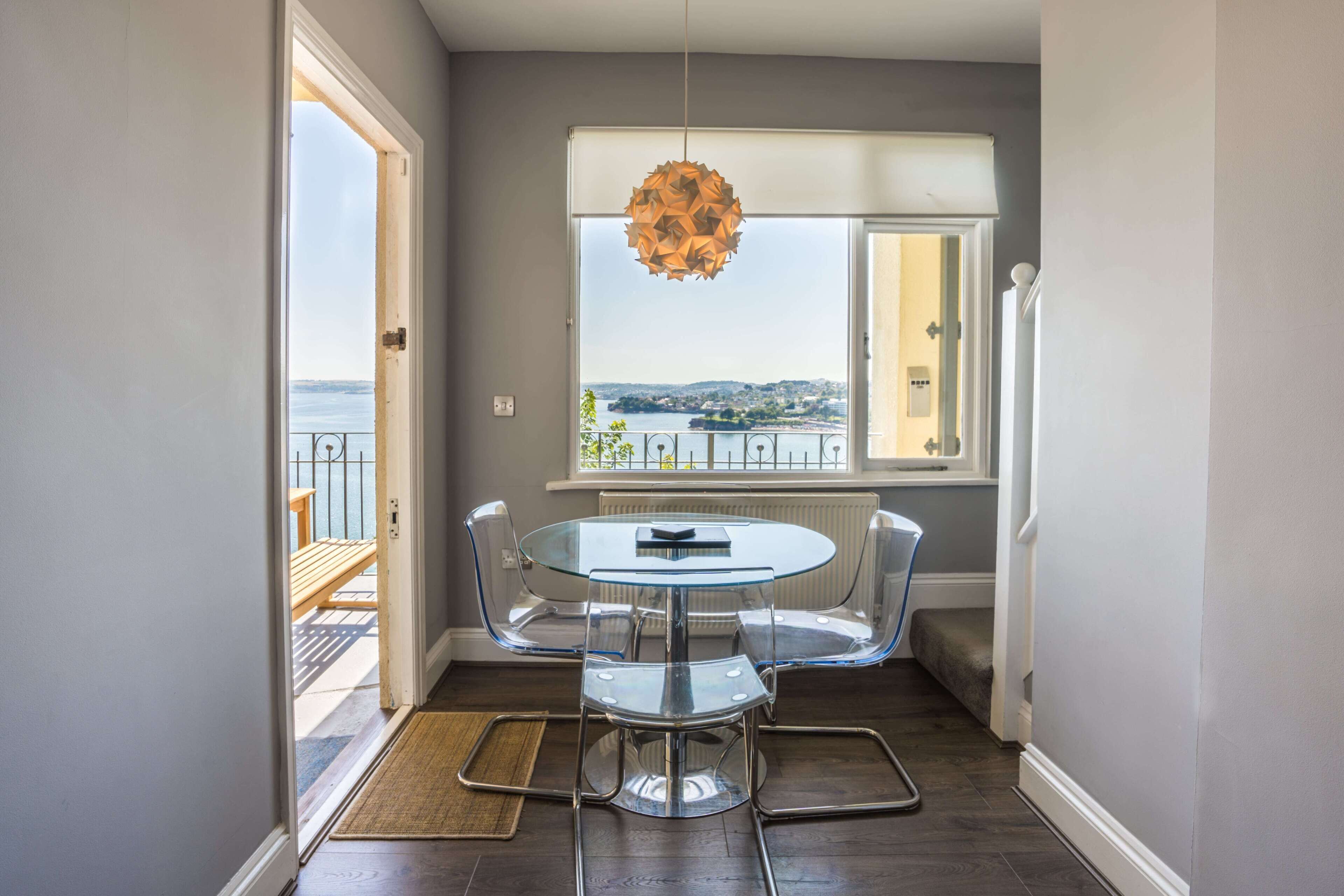 A dining area with a round glass table and chairs, a view of the sea through a window, and a hanging light fixture.