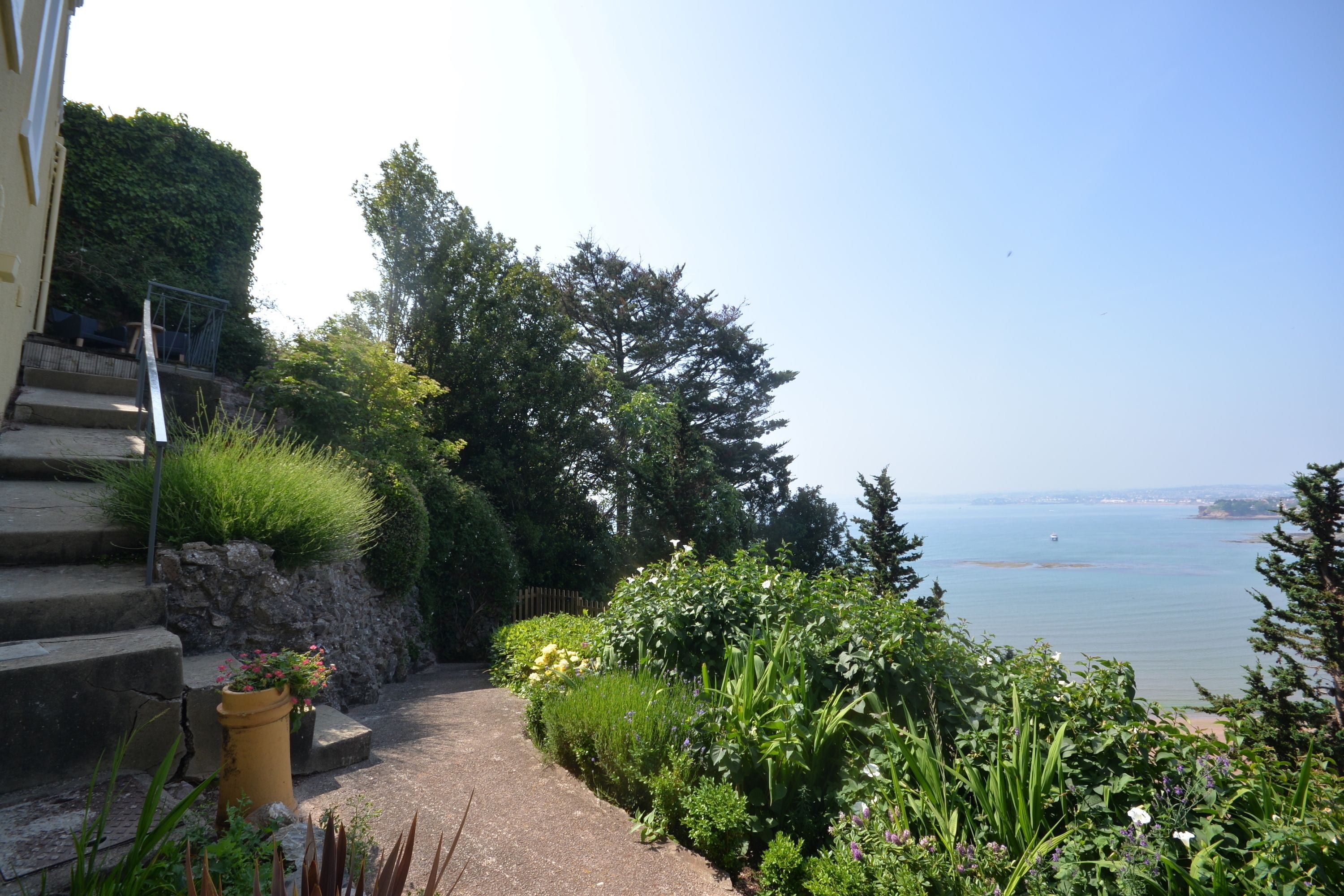 Un jardin en bord de mer, avec des marches menant à un chemin dallé, une végétation luxuriante et une vue sur la mer.