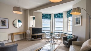 A modern living room with a glass coffee table, leather sofa, and a TV on a wooden stand. There are large windows with curtains, a round table, and a floor lamp. - Astor House (Torquay)
