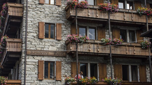 Un bâtiment en pierre doté de balcons en bois ornés de fleurs, une enseigne indiquant qu’il s’agit d’un hôtel, et une clôture en bois à l’avant.