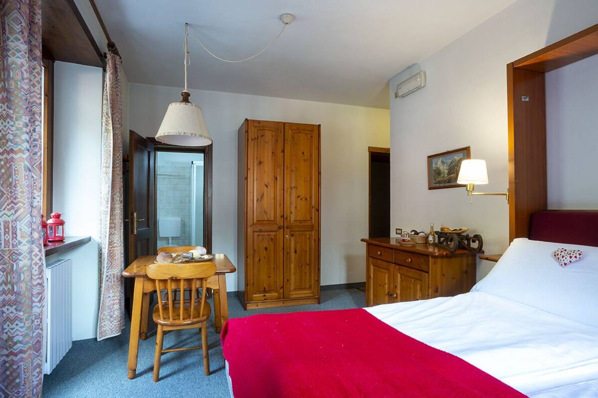 A bedroom with a bed, a wooden wardrobe, a desk with a chair, a hanging lamp, and a radiator.