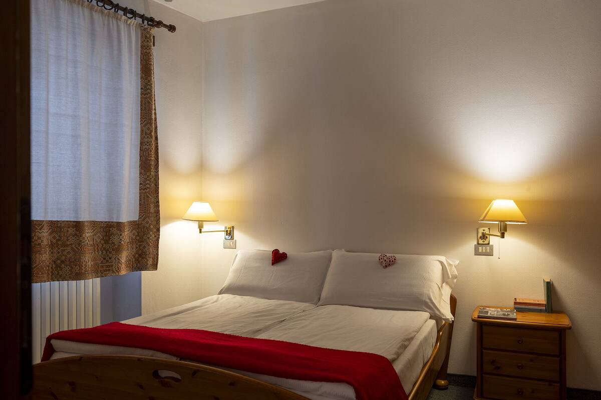 A neatly made bed with a red blanket, two bedside lamps, and a small wooden nightstand with a book.