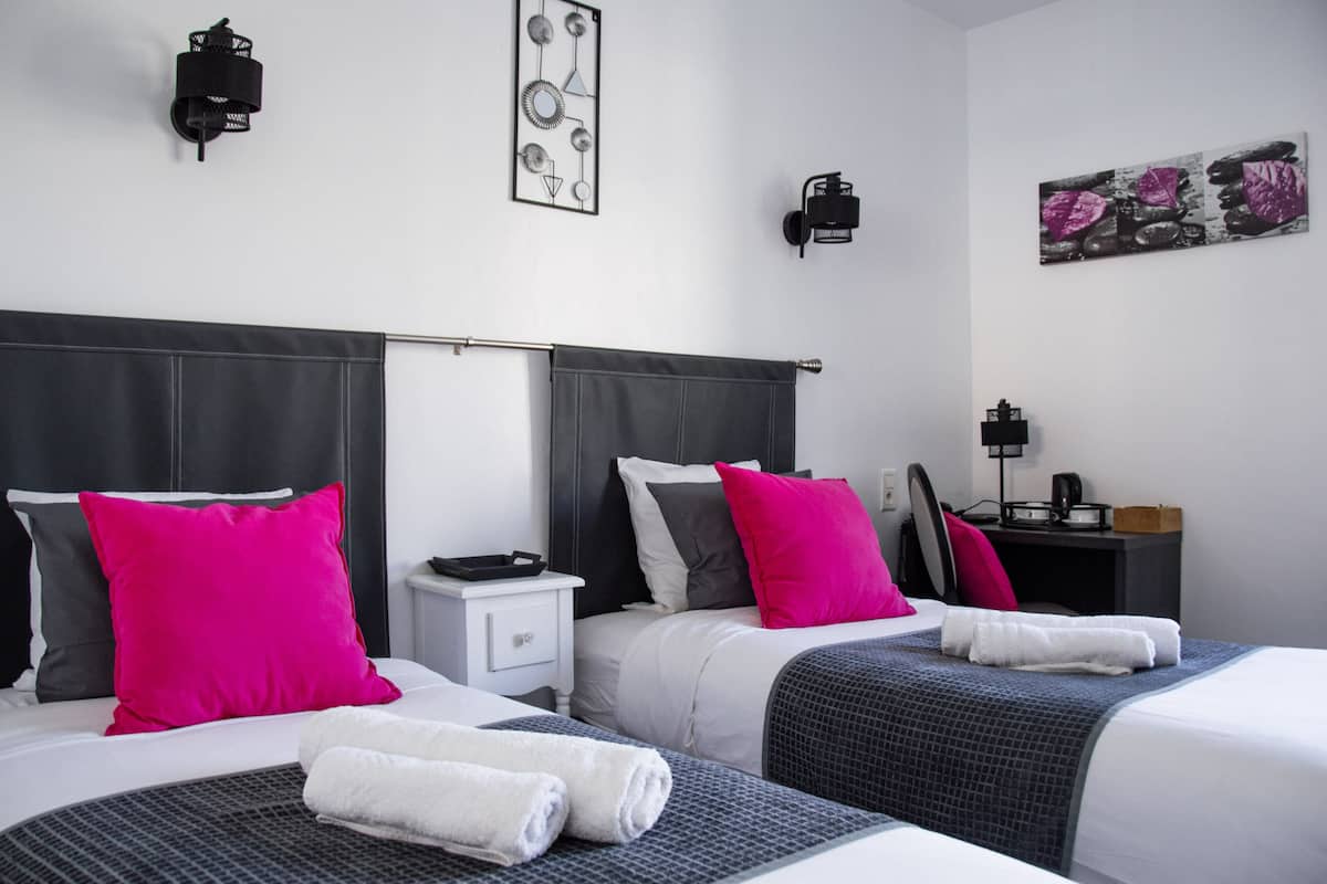 A hotel room with two beds, each with a headboard, white bedding, and bright pink pillows. There are two bedside tables, one with a lamp, and artwork on the walls.
