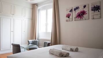 Habitación de hotel con una cama, una mesa de vidrio, dos sillas, un radiador y tres cuadros en la pared.