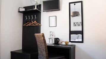 A hotel room with a dark wooden desk, a chair, a TV, a mirror, a wall-mounted fan, and a small kitchenette area.