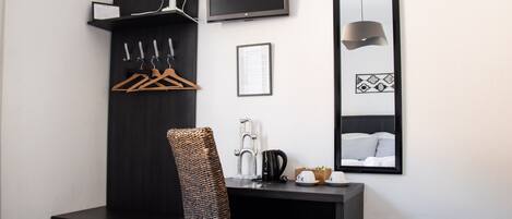 A hotel room with a dark wooden desk, a chair, a TV, a mirror, a wall-mounted fan, and a small kitchenette area.