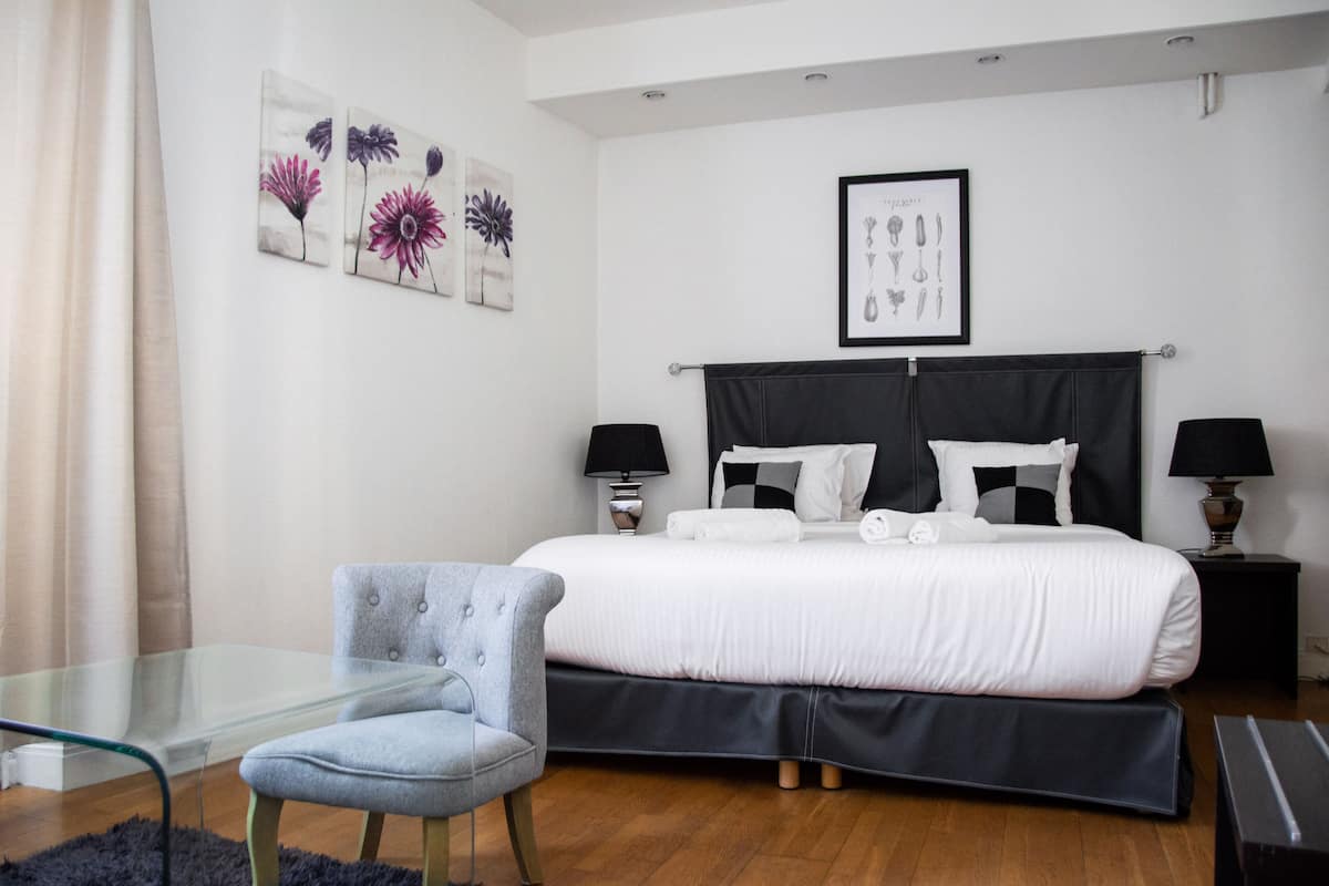 A modern bedroom with a large bed, two bedside tables with lamps, a glass table with a chair, and floral artwork on the wall.