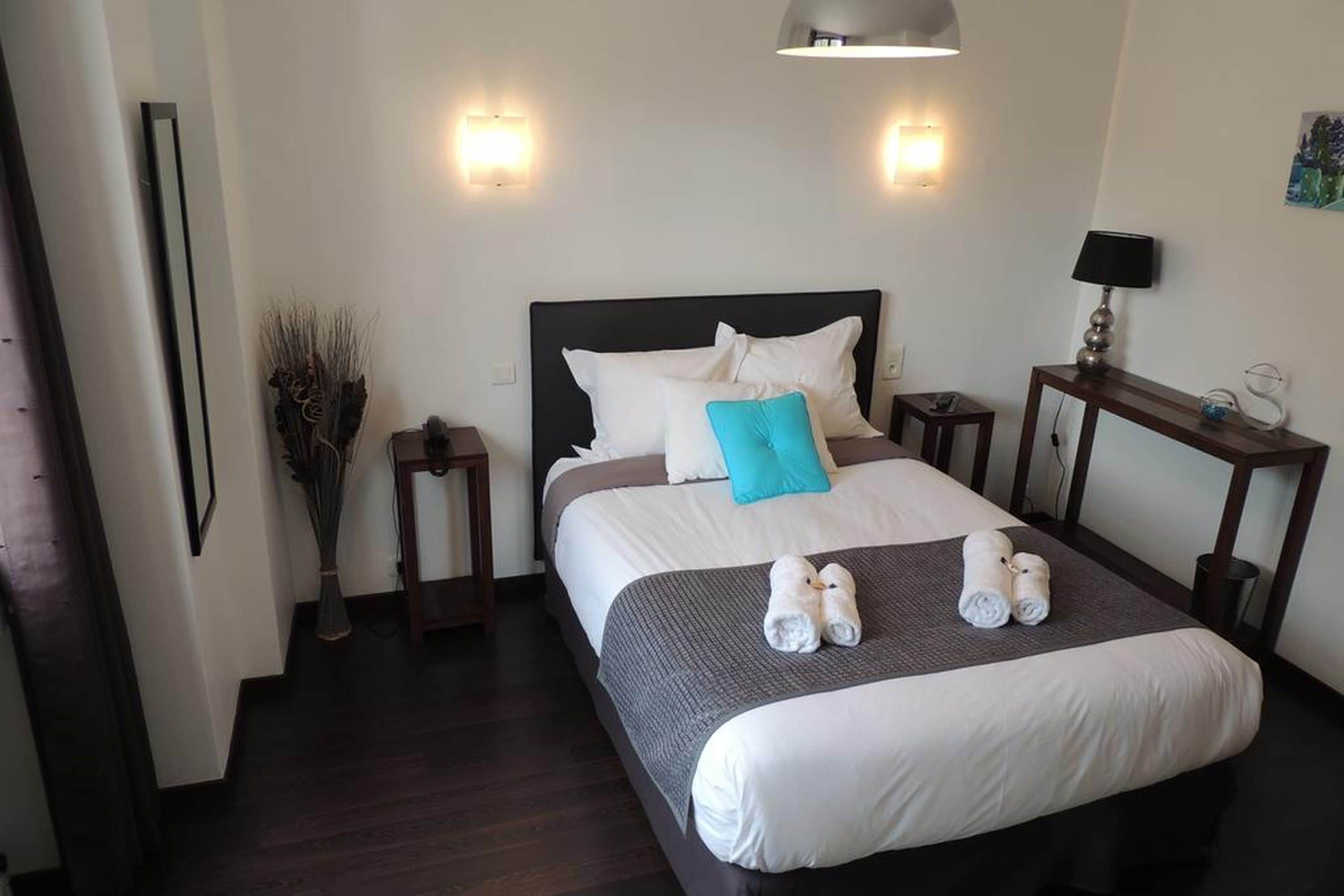 Una cama bien hecha con almohadas blancas y azules, dos mesitas de noche, una lámpara y un jarrón con plantas secas.