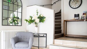 A modern hotel lobby with a grey armchair, a vase with white flowers, a black metal table, a staircase, and a clock on the wall.