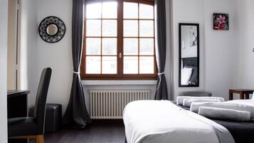 A hotel room with a large window, a bed with white linens, a chair, a small table, and a mirror on the wall.