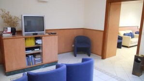 Una habitación con una televisión sobre un mueble de madera, sillas azules y una pequeña mesa.