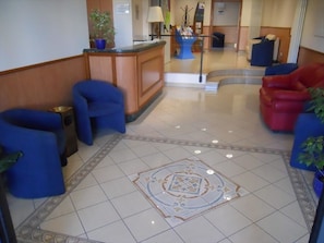 A lobby with a reception desk, blue and red seating, and a decorative floor pattern. - Hotel Italia (Ancona)