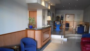 Un hall de hotel con mostrador de recepción, asientos azules y rojos, una escalera y una planta en maceta.