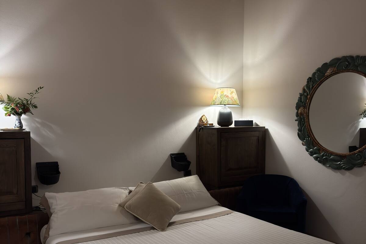 A bedroom with a bed, a nightstand, a lamp, a mirror, and a wall-mounted shelf.