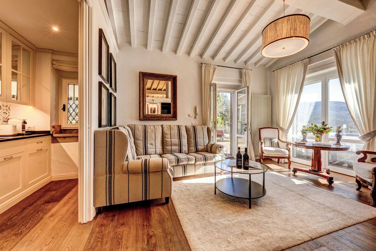 A spacious living room with a sofa, a coffee table, and a dining area with a table and chairs. There is a large window providing natural light and a view of the outdoors.