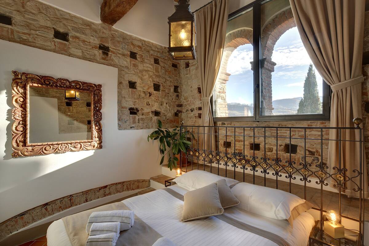 A bedroom with a stone wall, a large mirror, a bed with white linens, a potted plant, and a view through a window with curtains.