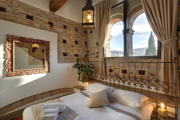 A bedroom with a stone wall, a large mirror, a bed with white linens, a potted plant, and a view through a window with curtains. - Terre di Baccio (Greve in Chianti)