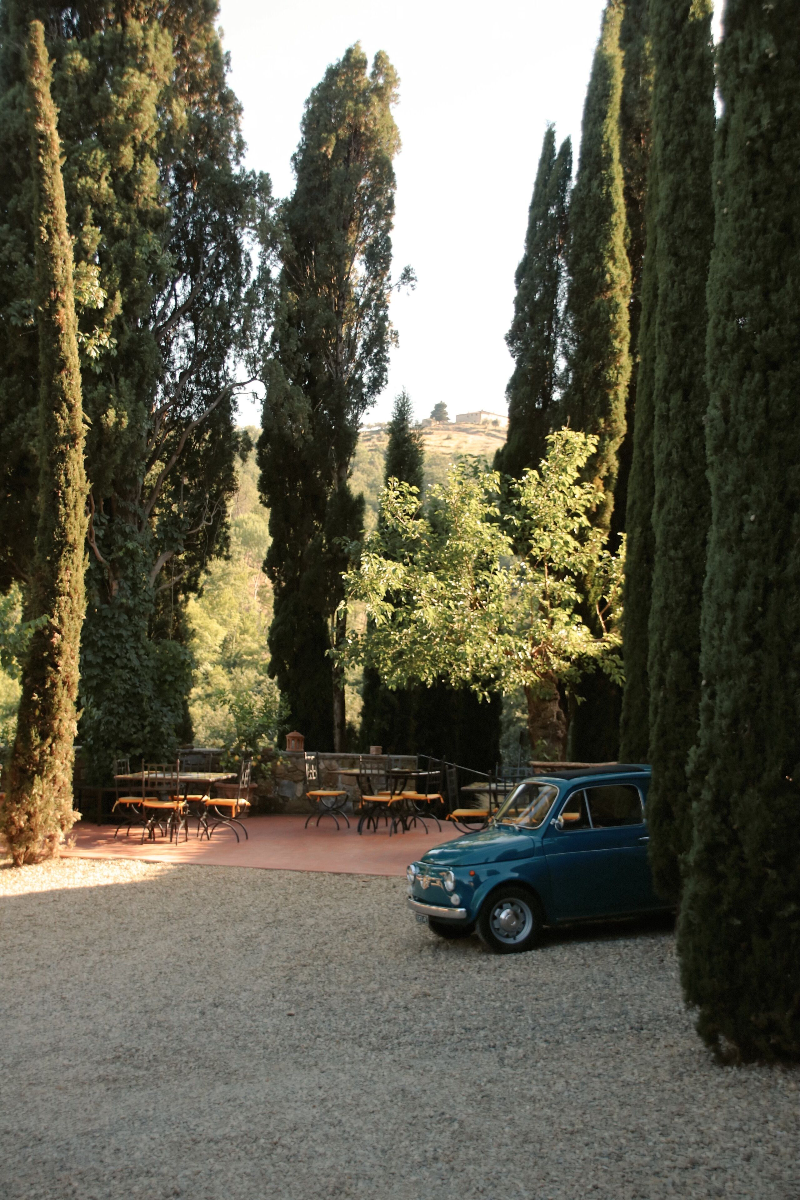 Un patio con altos cipreses, un coche azul antiguo y muebles de exterior.