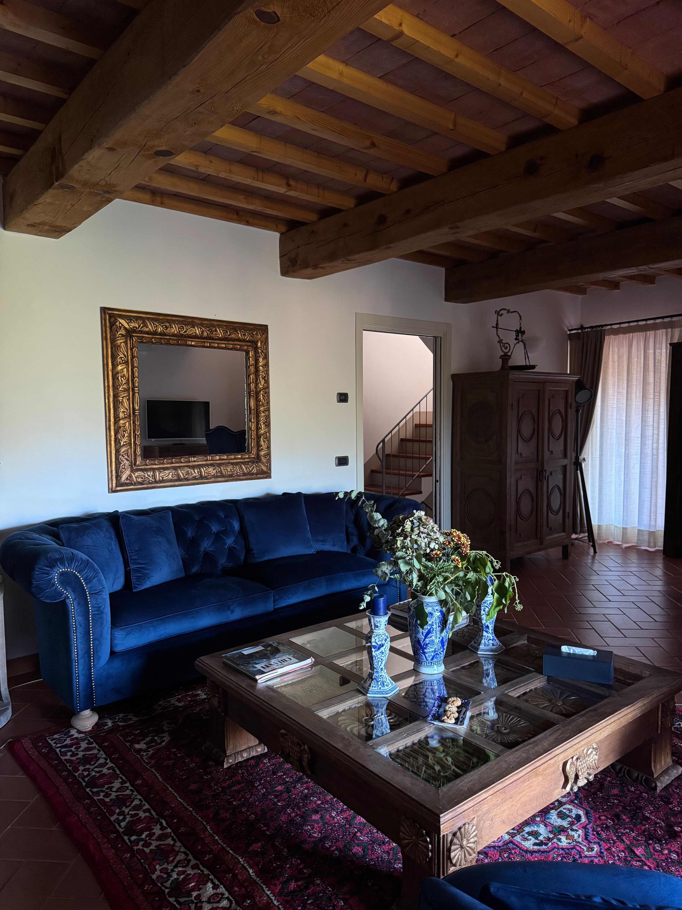 Una sala de estar con un sofá azul oscuro, una mesa de centro de vidrio con objetos decorativos, un techo de madera y un espejo grande en la pared.