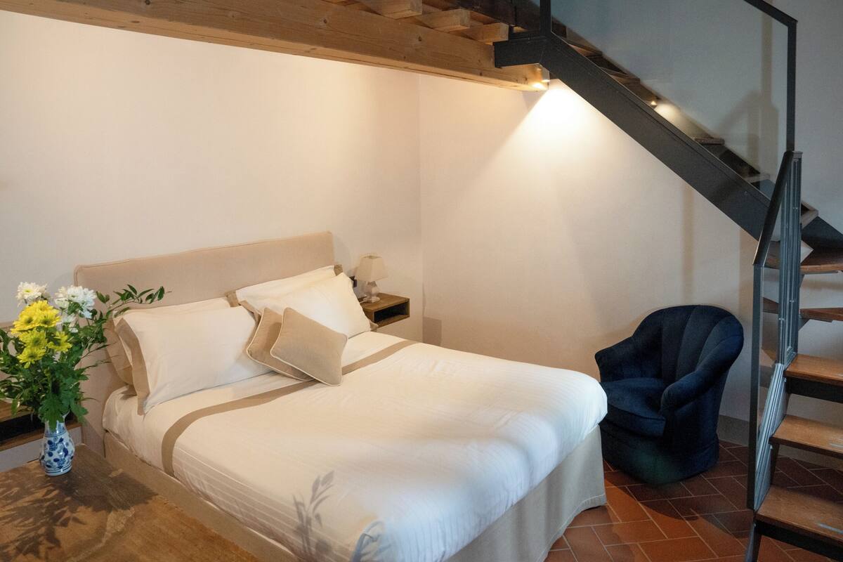 A bedroom with a bed, nightstand, chair, and a staircase.
