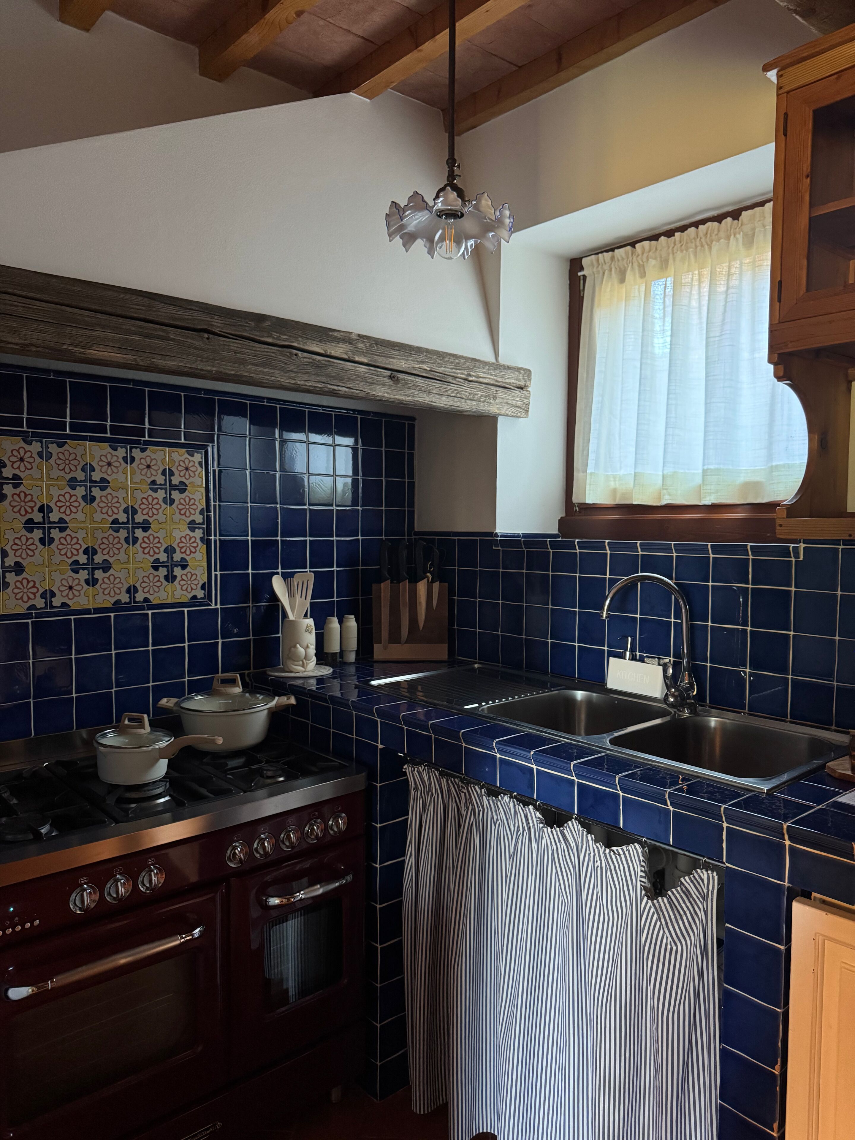 A kitchen with blue tiles, a stove, an oven, a sink, and a window with a curtain.