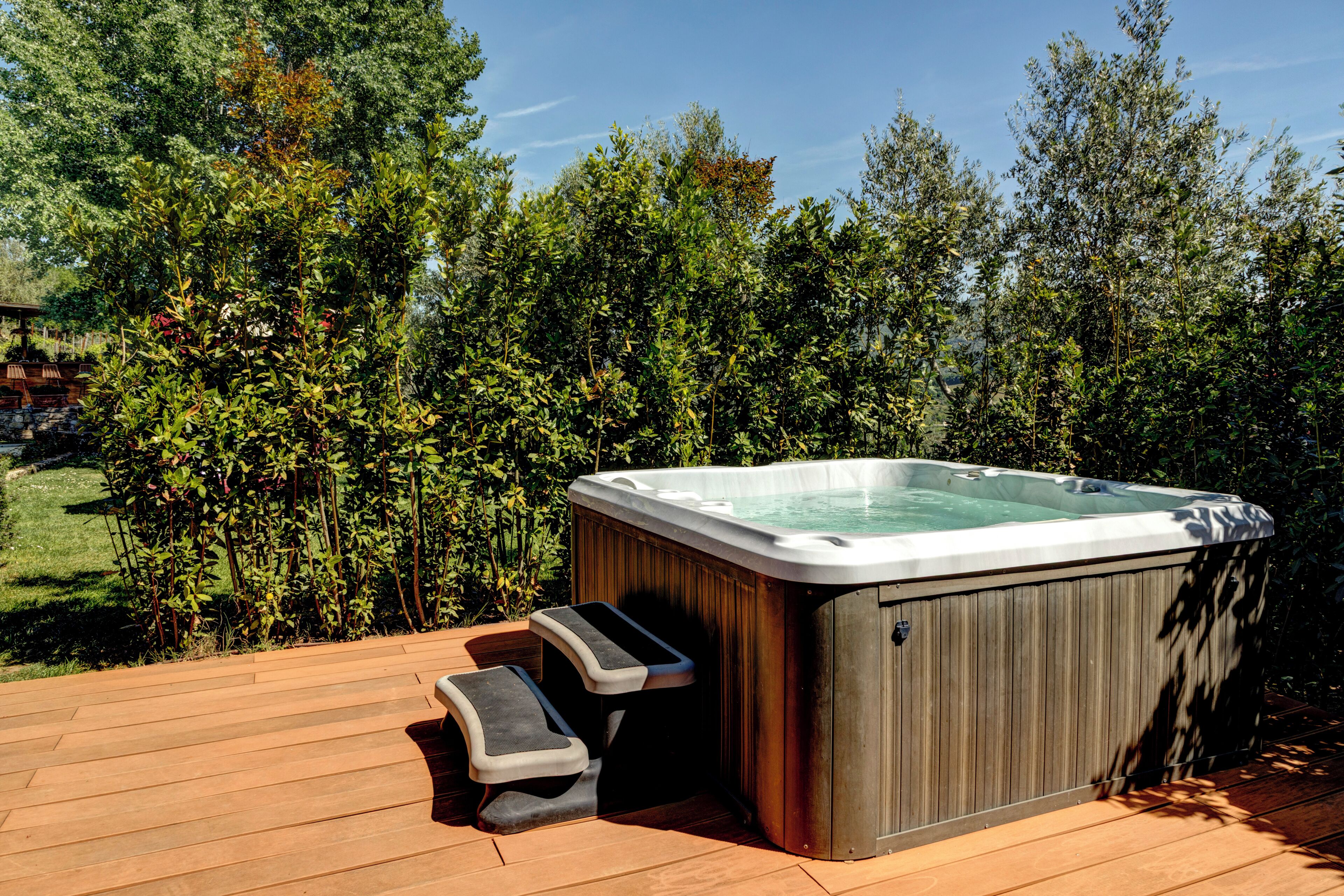 A hot tub on a wooden deck with steps leading up to it, surrounded by dense greenery.