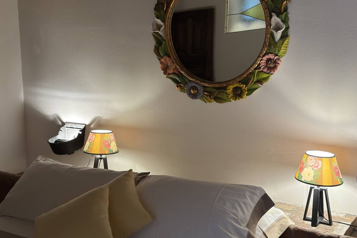A hotel room with a bed, two bedside tables with lamps, a wall-mounted mirror with a floral frame, and a small window.