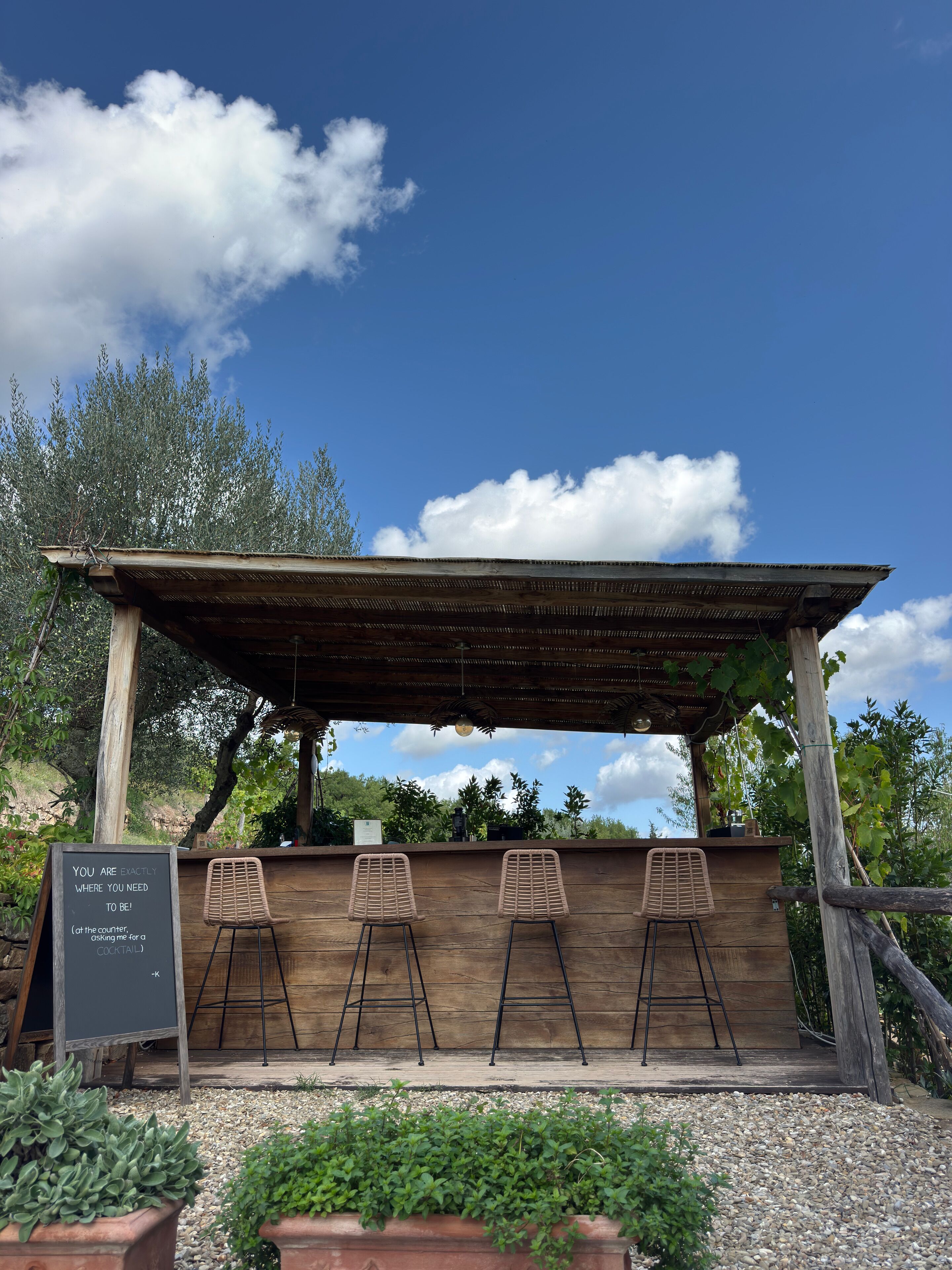A wooden outdoor bar with a pergola roof, three metal chairs, and a chalkboard sign.